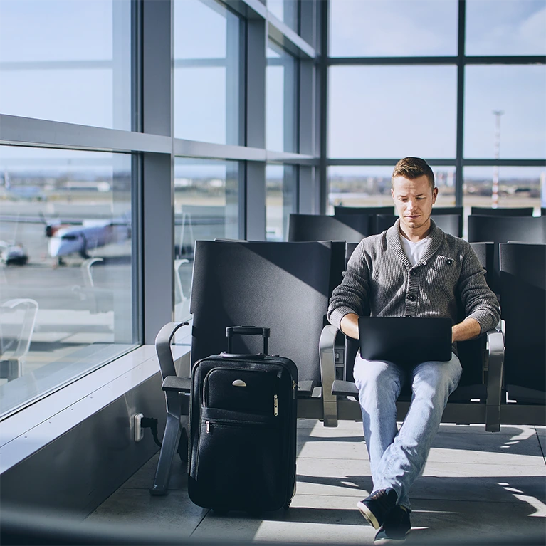 A man sits comfortably in the sunlight at an airport while working on his laptop.