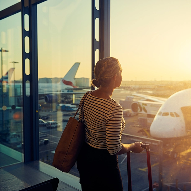 A woman stands in the airport looking out the window at the tarmac as the sun glares inside the terminal.