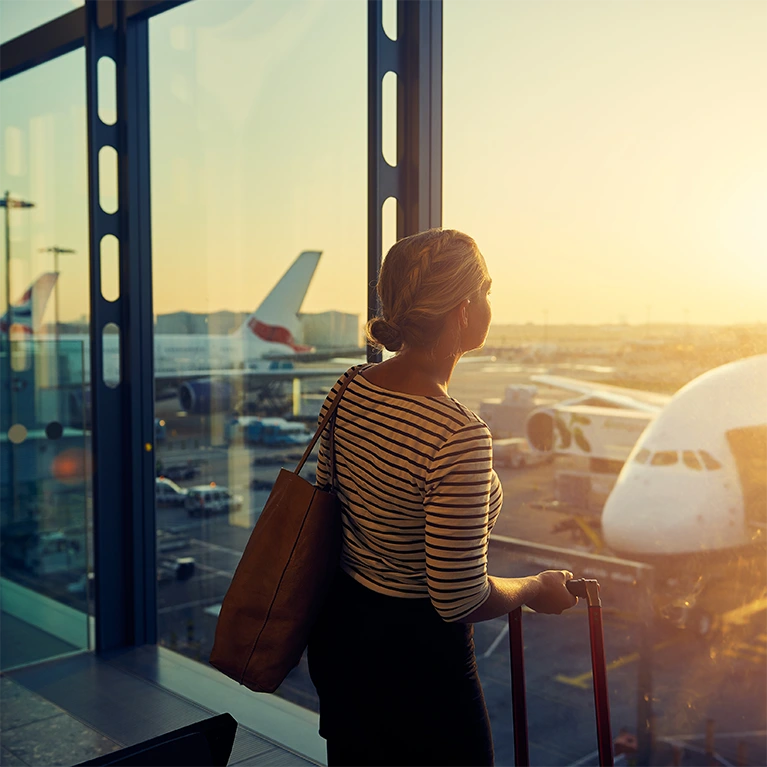 A woman stands in the airport looking out the window at the tarmac as the sun glares inside the terminal.