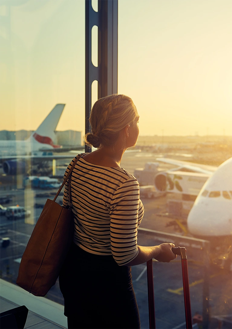 A woman stands in the airport looking out the window at the tarmac as the sun glares inside the terminal.
